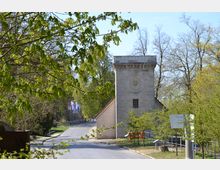 Steinturm mit einer Uhr auf der Fassade, umgeben von grünen Bäumen und einem mit Kopfstein gepflasterten Weg. Im Hintergrund sind Fahnenmasten und ein parkähnliches Gelände erkennbar.