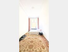 Schmales Schlafzimmer mit weiß gestrichenen Wänden, einem großen Teppich mit orientalischem Muster und einem niedrigen Bett vor einem Fenster mit rosafarbenen Vorhängen. Auf der rechten Seite steht ein Nachttisch mit einer Lampe, links ein Keyboard auf dem Boden.