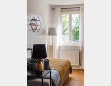Schlafzimmer mit einem Bett, das mit einer gelben Decke und grauer Bettwäsche bedeckt ist, einem Nachttisch mit Lampe und dekorativem Gegenstand, einem stehenden Lampenschirm, einem Heizkörper unter einem großen Fenster mit weißen Vorhängen und einer Wanddekoration mit geometrischen Regalen.