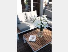 Holzterrasse mit modernen Gartenmöbeln, darunter eine weiße Sitzgruppe mit grauen Polstern und einem rechteckigen Couchtisch aus Holz. Auf dem Tisch stehen eine Vase mit weißen Lilien und ein Buch mit einem Porträt auf dem Cover.