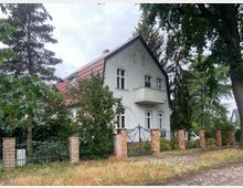 Zweistöckiges Wohnhaus mit weißer Fassade, rotem Satteldach und einem kleinen Balkon. Das Grundstück ist von einer gemauerten Zaunbegrenzung mit Metalltor umgeben, umgeben von hohen Bäumen und Sträuchern.