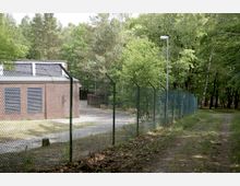 Backsteinbau mit vergitterten Fenstern, umgeben von einem hohen Maschendrahtzaun mit Stacheldraht; im Hintergrund dichter Wald und eine Nebenstraße, beleuchtet von einer Straßenlaterne.