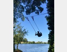 Ein Mensch schaukelt an einer Schaukel, die zwischen Bäumen aufgehängt ist, mit Blick auf einen See. Im Hintergrund sind grüne Ufer und ein klarer, blauer Himmel zu sehen.