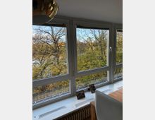 Innenaufnahme eines hellen Raumes mit großen, rechteckigen Fenstern, die auf eine herbstliche Baumlandschaft mit gelblichen Blättern blicken. Auf der weißen Fensterbank stehen kleine gerahmte Fotos, darunter befindet sich ein Heizkörper, und im Vordergrund sind ein Holztisch und ein weißer Stuhl sichtbar.