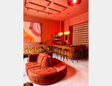 Modern lounge bar interior with red walls, a curved rust-colored sectional sofa, and a wooden bar lined with tall black stools under hanging globe pendant lights. The space includes an abstract wall painting, a small back bar shelf, and a patterned brick screen along one side.
