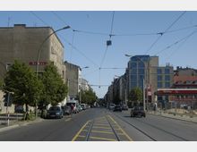 Straßenszene in einer Stadt mit zweispuriger Fahrbahn, Straßenbahnlinien in der Mitte und beidseitig parkenden Autos. Mehrstöckige Gebäude, darunter ein moderner Glasbau und ein Tankstellengebäude, säumen die Straße; im Vordergrund stehen Straßenlaternen und Bäume.