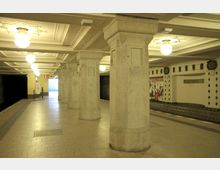 U-Bahn-Station mit symmetrischen, massiven Säulen aus Stein, abgehängten Decken mit integrierten Leuchten und einem Bahnsteig aus Fliesen. Im Hintergrund sind Stufen, eine Wand mit Wandgestaltung und ein Schild mit der Aufschrift „Rüdesheimer Platz“ zu sehen.