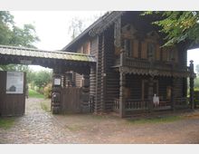 Holzgebäude im russischen Stil mit Veranda und ornamentalem Holzschnitt an Balken und Geländern, daneben ein hölzernes Tor mit einem Schild, das den Eingang zu einem Museum markiert; Kopfsteinpflasterweg und Garten im Hintergrund.