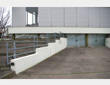 Modernes Gebäude mit einer Fassade aus grauen Lamellen und verglasten Eingängen im Untergeschoss. Im Vordergrund sind ein Betonweg, Treppen, weiße Abgrenzungsmauern und Metallgeländer zu sehen.