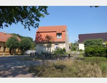 Ein kleines Wohnhaus mit rotem Ziegeldach und verblasster Fassade, umgeben von einem Garten und niedrigen Zaun. Links und rechts befinden sich weitere Gebäude mit ähnlich gestalteten Dächern, eingebettet in eine ländliche Umgebung mit Bäumen und Sträuchern.