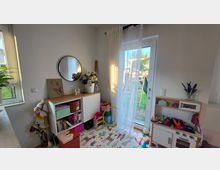 Heller Spielbereich in einem Wohnraum mit weißer Wand, niedrigem Regal mit rundem Spiegel darüber und Spielteppich auf dem Boden. Rechts führt eine verglaste Terrassentür mit transparenten Vorhängen in den Garten; daneben steht eine Kinder-Spielküche und verschiedene Spielsachen.