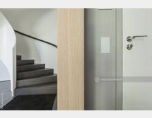 Moderner Innenbereich mit einer Holzsäule im Vordergrund, einer weißen Tür mit Metallklinke rechts und einer dunkel gehaltenen Wendeltreppe links, die von einer weißen Wand umgeben ist. Der Boden ist mit einem dunklen Teppich und grauen Fußboden ausgestattet.