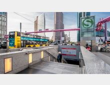 Straßenszene am Potsdamer Platz in Berlin mit modernen Hochhäusern im Hintergrund, einem gelben Doppeldeckerbus im Vordergrund und einem Zugang zur S-Bahn-Station mit grünem "S"-Symbol. Auffällige rosa Rohre und Hinweisschilder ergänzen das Stadtbild.