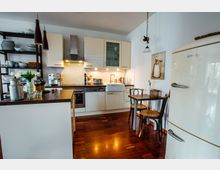 Moderne Küche mit weißen Einbauschränken, einer Arbeitsplatte aus Holz und Geräten wie einem Ofen, einer Dunstabzugshaube und einer Kaffeemaschine. Im Vordergrund steht ein kleiner Esstisch mit zwei Holzstühlen, daneben ein cremefarbener, retro-stiliger Kühlschrank.