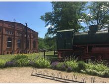 Historisches Backsteingebäude mit großen Fenstern auf der linken Seite, daneben eine grüne Dampflokomotive hinter einem Zaun. Im Vordergrund befinden sich Blumenbeete und ein Fahrradständer, umgeben von Bäumen und blauem Himmel im Hintergrund.