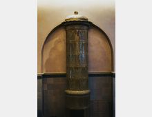 Historischer Innenraum mit einem hohen, zylindrischen Kachelofen in einer Wandnische, verziert mit reliefierten Blattmotiven und einer gewölbten Ofenhaube. Die Wand ist zweifarbig gehalten, unten mit Fliesen und oben mit glattem Putz.