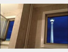 Blick aus einem Innenraum mit weiß gerahmten Fenstern; durch eines der Fenster ist der Berliner Fernsehturm bei Nacht vor dunklem Himmel zu sehen. Die Wände sind schlicht mit sichtbaren Rohren.