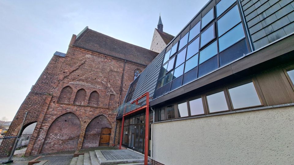 Außenansicht eines Backstein-Kirchengebäudes mit spitzem Turm, neben dem ein moderner Anbau mit großen Glasfenstern und schräger Metallverkleidung anschließt. Im Vordergrund führen Stufen zu einem verglasten Eingangsbereich; die Kirchenwand zeigt gemauerte Rundbogen-Nischen und eine zugemauerte Türöffnung.
