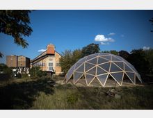 Historisches Industriegebäude aus Backstein mit großen Fenstern und Giebeldach, daneben eine transparente Kuppelstruktur aus Holzstreben auf einer freien Fläche, umgeben von Bäumen und Wiese unter blauem Himmel.