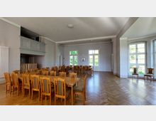 Großer, heller Raum mit hölzernem Parkettboden und mehreren Holztischen und Stühlen, die in Reihen angeordnet sind. Große Fenster und eine Glastür lassen viel Tageslicht herein, während an einer Wand eine kleine Empore und Heizkörper zu sehen sind.