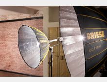 Zwei große Studioleuchten mit reflektierenden, silbernen Innenseiten und schwarzer Außenseite mit der goldenen Aufschrift "BRIESE Focus 220-H" stehen in einem Raum mit einer Backsteinwand und einer Metalltür im Hintergrund.