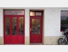 Fassade eines Gebäudes mit weißer Putzwand und roten Türen und Fenstern. Links befindet sich ein Schaufenster mit der Aufschrift „Location Network“ und rechts ein Motorrad, das teilweise sichtbar ist.