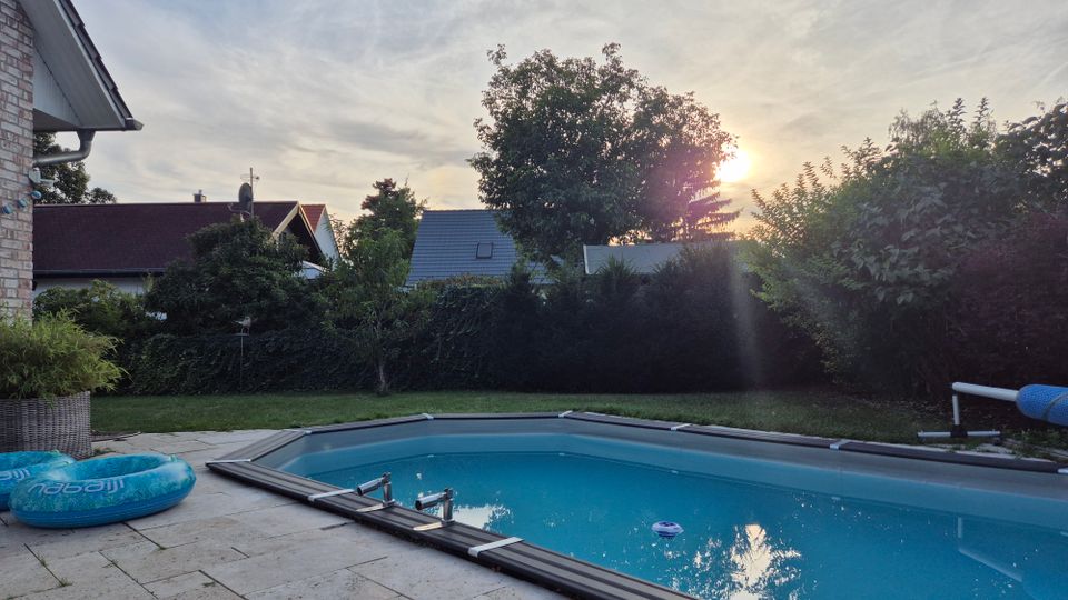 Gartenansicht mit rechteckigem Swimmingpool, umgeben von einer gefliesten Terrasse und Rasenfläche; im Hintergrund sind Hecken, Bäume und Wohnhäuser mit Satteldächern sowie ein Sonnenuntergang zu sehen.