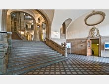 Großes Foyer eines historischen Gebäudes mit einer breiten Steintreppe, geschwungenen Rundbögen, verzierten Wandreliefs und hohen Decken. Der Boden ist mit einem gemusterten Fliesenmuster gestaltet, während auf der rechten Seite eine offene Holztür und Informationsschilder zu sehen sind.