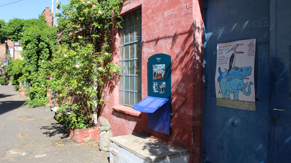 Außenansicht eines niedrigen Backsteingebäudes mit großem Fenster und blauer Tür, an der „Theater im Bergmannkiez“ steht. Vor der Fassade rankt eine blühende Kletterpflanze, daneben befindet sich eine Informationstafel und ein Plakat, entlang eines gepflasterten Hofwegs.