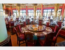 Großes, elegant dekoriertes chinesisches Restaurant mit roten Wänden, vergoldeten Säulen und floralen Schnitzereien auf Glasflächen. Im Vordergrund steht ein runder, glänzender Holztisch mit roten Polsterstühlen und gedeckten Plätzen.