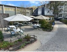 Außenbereich eines kleinen Cafés mit gepflastertem Boden, weißen Tischen, grünen Stühlen und weißen Sonnenschirmen. Im Hintergrund steht ein kleines, historisches Gebäude mit Schieferdach und Fachwerkelementen, umgeben von Pflanzen und Bäumen.