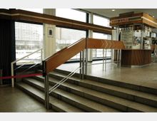 Innenraum eines Gebäudes mit einer Treppe und einer Rampe mit Holzgeländer, daneben ein Kassenhäuschen mit Holzverkleidung und einem digitalen Schriftzug. Große Fenster bieten Blick auf ein benachbartes, mehrstöckiges Gebäude.