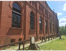 Ziegelsteinfassade eines historischen Industriegebäudes mit großen, hohen Fenstern und Rundbögen. Im Vordergrund eine schmale Metallkonstruktion und Baumstümpfe auf einer Rasenfläche; im Hintergrund ein klarer Himmel mit einigen Wolken.