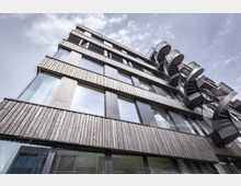 Moderne mehrstöckige Gebäudefassade mit einer vertikalen Holzverkleidung und großen Glasfenstern; rechts sind markante, spiralförmige Außentreppen aus Metall angebracht. Der Himmel ist leicht bewölkt und bildet den Hintergrund.