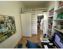 Zimmer in einer Wohnung mit Holzboden und hohen Decken, eingerichtet als Arbeitsbereich. Zu sehen sind ein Schreibtisch mit Computer, ein Bücherregal, eine Collage an der Wand, ein Schrank, sowie ein Hochbett über einer Zugangstür.