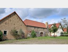 Backsteingebäude mit roten Ziegeldächern auf einem ländlichen Grundstück, umgeben von Bäumen und Sträuchern. Im Vordergrund eine Wiese, im Hintergrund weitere Gebäude mit weißer Fassade und einem kleinen Gartenbereich.