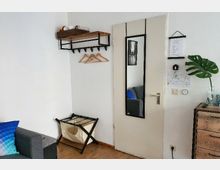 Ein kleiner, funktionaler Raum mit einer weißen Tür, an der ein schmaler Spiegel hängt. An der Wand befindet sich eine Garderobe mit Holzablage und Kleiderhaken, daneben eine Kommode mit einer grünen Monstera-Pflanze in einer Vase.