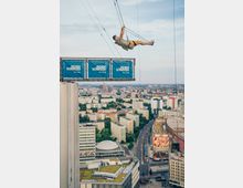 Ein Mann hängt an einem Seil in der Luft, beim sogenannten Base Flying von einer Plattform mit dem Schriftzug „Jochen Schweizer“ über einer Stadtlandschaft. Im Hintergrund sind moderne Gebäude mit Flachdächern, Hochhäuser, Straßen und Grünflächen zu sehen.