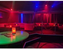 Gedimmter Lounge- oder Clubinnenraum mit roten und blauen Deckenlichtern, runden Tischen mit Kerzenlampen und umlaufenden Polstersofas. Vor einer erhöhten Sitzfläche mit Seilabsperrung hängen dunkle Vorhänge an der Rückwand.
