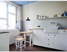 Ein schlichtes, helles Kücheninterieur mit weißen Schränken, einer tiefen Spülbeckenarmatur und einer wandmontierten Ablage mit Gefäßen. Links befindet sich ein großes Fenster mit weißem Rahmen, darunter ein kleiner Esstisch mit Holzoberfläche und zwei passenden Hockern.