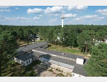 Eine von Wald umgebene Anlage mit einstöckigen, flachen Gebäuden und asphaltierten Wegen, im Hintergrund ein weißer Aussichtsturm und Windräder sichtbar unter einem blauen Himmel mit Wolken.