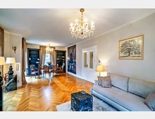 Elegant eingerichtetes Wohnzimmer mit Parkettboden, hellen Wänden und einem Kristallleuchter an der Decke. Im Raum befinden sich eine Sitzecke mit Sofa, ein schwarz gemusterter Couchtisch, ein Esstisch mit gepolsterten Stühlen vor Bücherregalen, sowie Wanddekorationen wie ein Gemälde und ein Baumdruck.