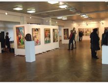 Ein moderner Ausstellungsraum mit Gemälden an weißen Stellwänden und auf Sockeln präsentierten Skulpturen. Der Raum hat einen Holzboden, eine Decke mit integrierter Beleuchtung, und einige Besucher betrachten die Kunstwerke.