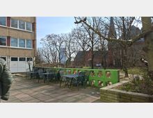 Gepflasterte Außenterrasse neben einem mehrstöckigen Gebäude mit mehreren Metalltischen und -stühlen sowie einer niedrigen grünen Betonbrüstung mit ovalen Öffnungen. Dahinter liegen ein kleiner Grünbereich mit kahlen Bäumen und im Hintergrund ein rotes Backsteingebäude mit Kirchturm.