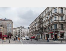 Straßenszene in einer Stadt mit mehrstöckigen Altbauten im Gründerzeitstil, die Läden im Erdgeschoss beherbergen, darunter ein Bistro und ein Grillhaus. Im Vordergrund sind Fußgänger, Radfahrer und Autos an einer Kreuzung mit Ampel zu sehen, während der Himmel bewölkt ist.