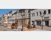 Straßenszene in einer Fußgängerzone mit historischem Charakter; links und rechts stehen mehrstöckige Gebäude mit beige- und pastellfarbenen Fassaden. Im Vordergrund befinden sich stilisierte Kunst- und Sitzobjekte aus Ziegelsteinen, sowie moderne Straßenlaternen.