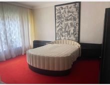 Schlafzimmer mit rotem Teppichboden, rundem Bett mit gepolstertem Rahmen in beigem Stoff und einer großen, schwarz-weißen Kunstdarstellung an der Wand. Links ist ein bodentiefes Fenster mit transparenten Vorhängen sichtbar.