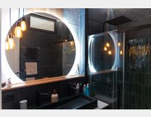 Modernes Badezimmer mit dunklen Fliesenwänden und Holzdetails. Ein großer runder Spiegel mit Beleuchtung, ein Waschbecken mit schwarzer Armatur und eine Glasdusche sind sichtbar; daneben hängen mehrere leuchtende Glühbirnen im Vintage-Stil.