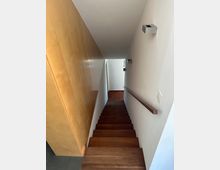 Blick in ein schmales, innenliegendes Treppenhaus mit dunklen Holzstufen, weiß gestrichenen Wänden und Handlauf rechts. Links verläuft eine große Holzverkleidung, am Ende führt die Treppe zu einem kleinen Flur mit Holzfußboden.