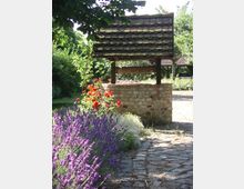 Brunnen mit einem Dach aus Holzschindeln, umgeben von einem gepflasterten Weg und Blumenbeeten mit blühendem Lavendel und roten Blumen, in einem ruhigen Garten mit Bäumen im Hintergrund.